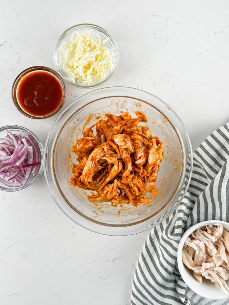 A glass bowl of shredded chicken mixed with BBQ sauce sits on a white surface, surrounded by small bowls of barbecue sauce, shredded cheese, sliced red onion, and plain shredded chicken. A striped cloth is nearby.