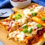 Rectangular slices of barbecue chicken pizza topped with melted cheese, red onions, and chopped green onions sit on a wooden board. A bowl of barbecue sauce and a pizza cutter are in the background.