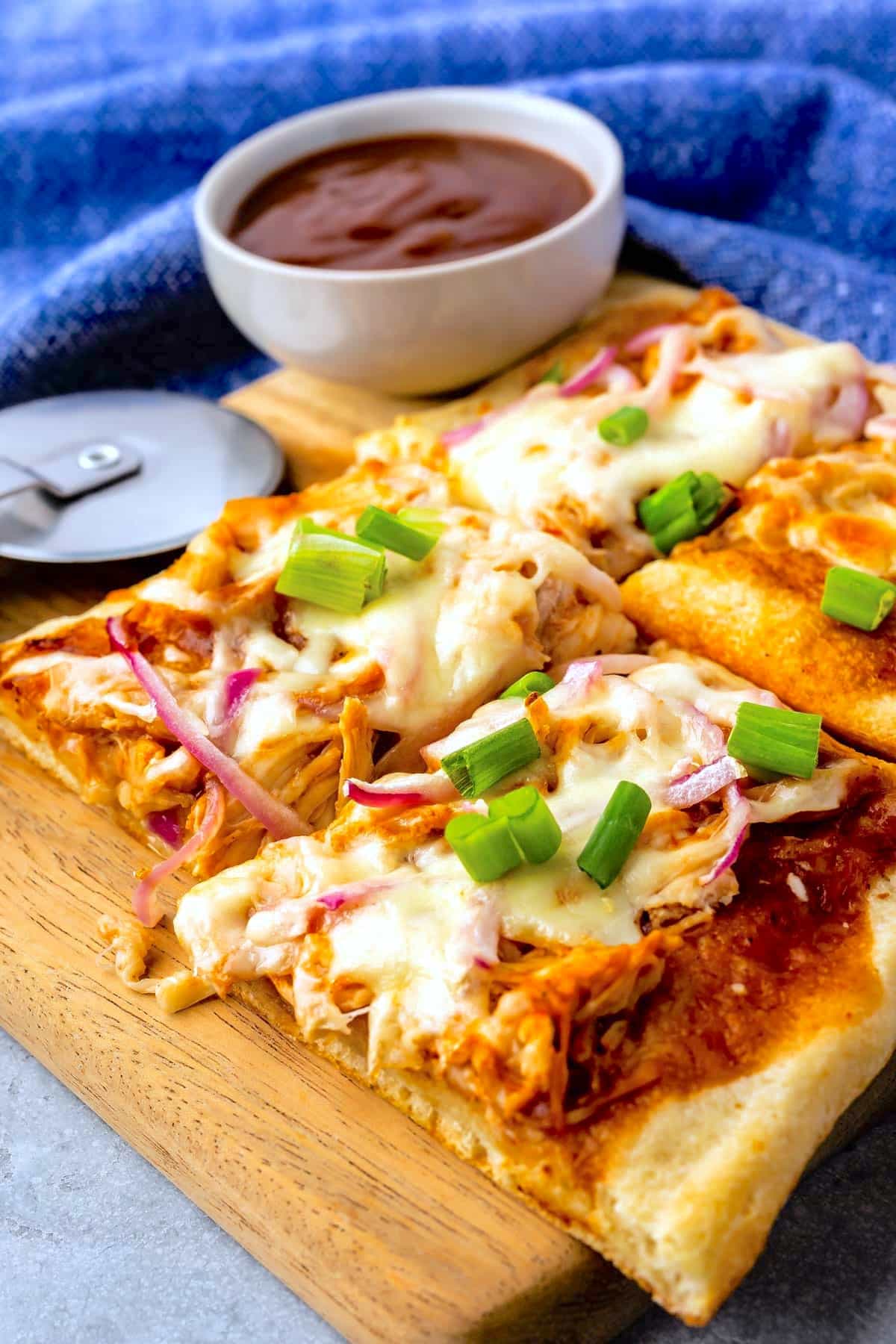 Rectangular slices of barbecue chicken pizza topped with melted cheese, red onions, and chopped green onions sit on a wooden board. A bowl of barbecue sauce and a pizza cutter are in the background.