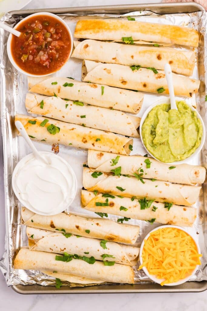 A tray of baked taquitos on foil, garnished with chopped herbs. Surrounding the taquitos are small bowls of salsa, guacamole, shredded cheddar cheese, and sour cream. Spoons are in the salsa, guacamole, and sour cream.