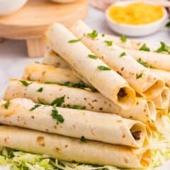 A plate of rolled and baked taquitos filled with meat is served on a bed of shredded lettuce, garnished with chopped parsley. Small bowls of salsa, sour cream, guacamole, and shredded cheese are in the background.