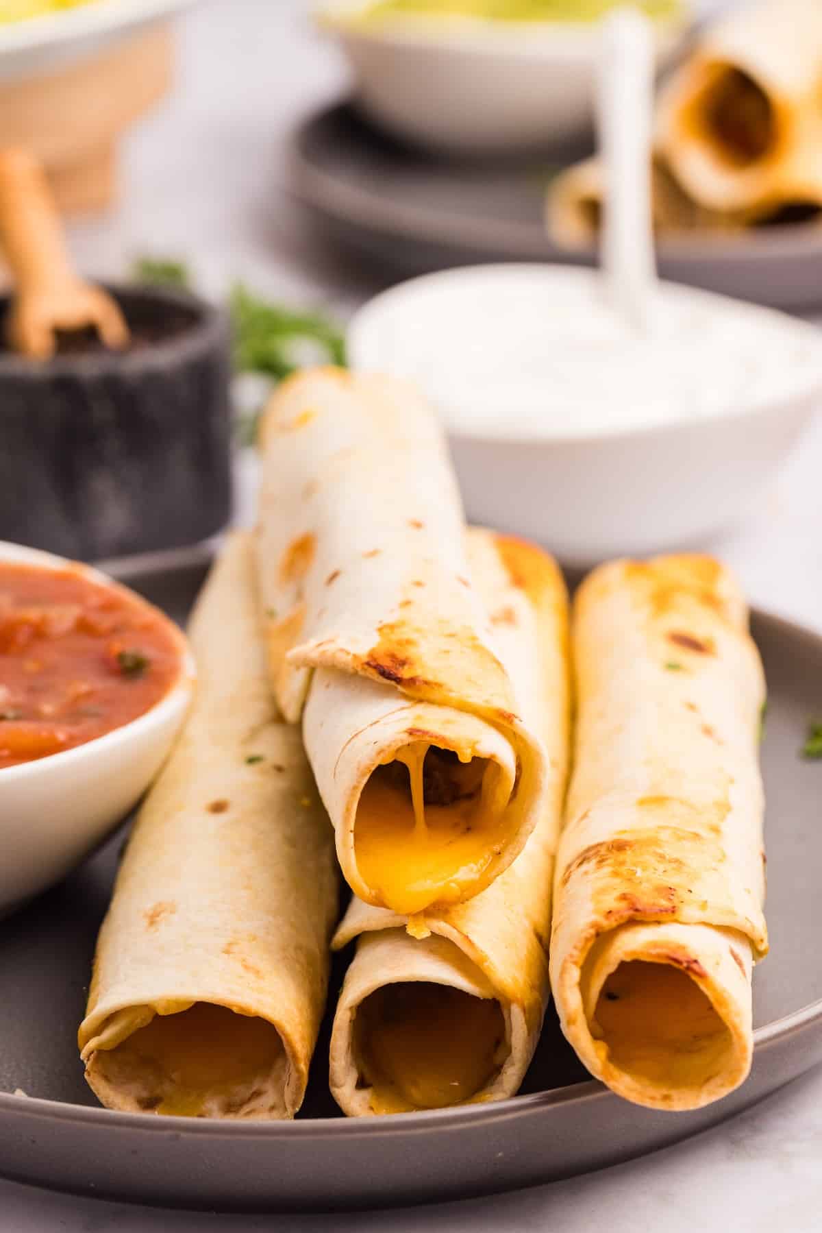 Four baked taquitos with melted cheese filling are stacked on a plate. A bowl of salsa and another bowl with a white creamy dip sit nearby. The background shows more food and a blurred dining setting.