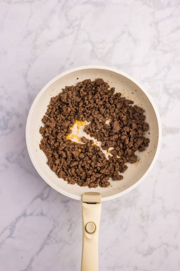 A white frying pan with cooked ground beef sits on a light marble countertop. The beef is browned and crumbled, with a small pool of grease visible in the center.