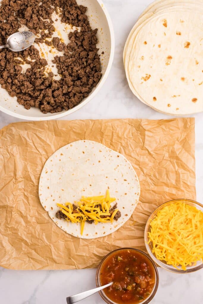 A tortilla on brown paper is topped with ground beef and shredded cheese. Nearby are a bowl of salsa with a spoon, a bowl of shredded cheese, a skillet of cooked beef, and a stack of tortillas.