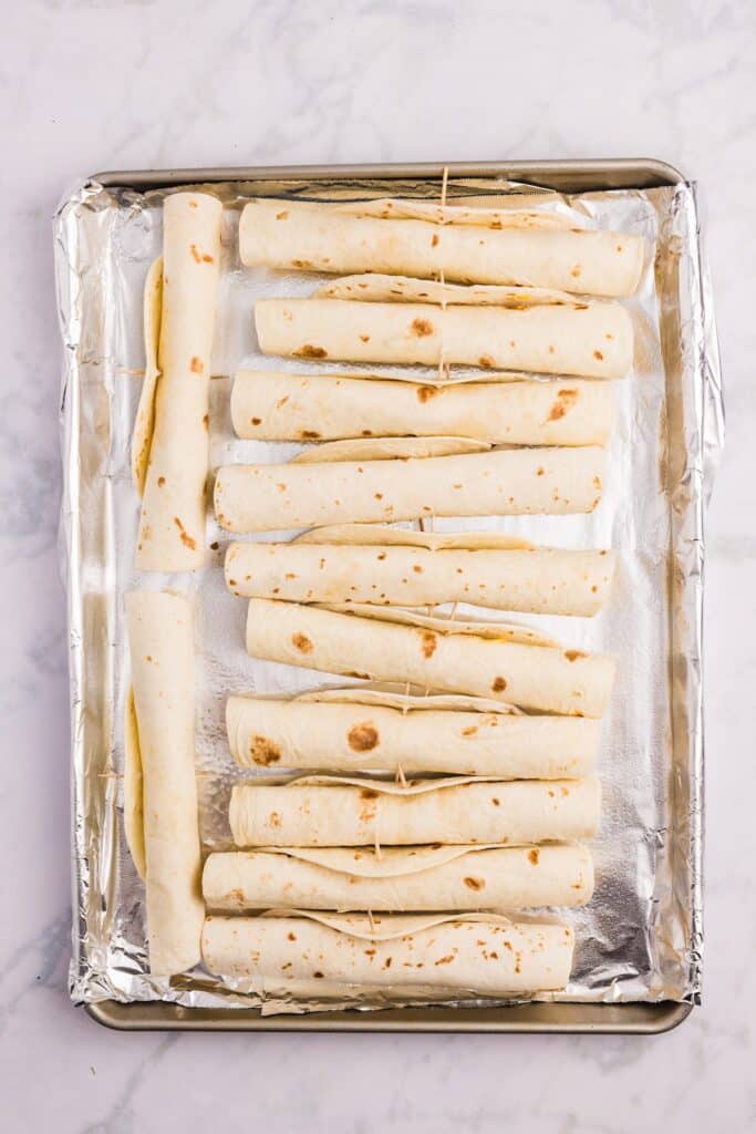 A baking sheet lined with foil holds several rolled tortillas, each secured with a toothpick, arranged neatly in rows on a marble surface.