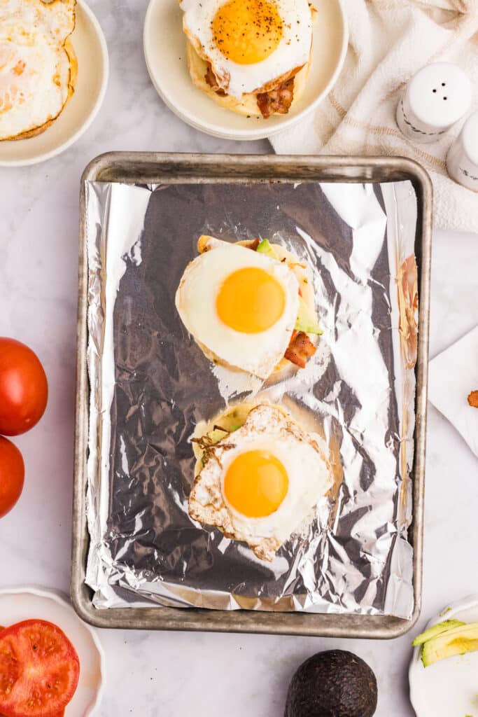 An open-faced breakfast sandwich topped with a sunny-side-up egg, avocado slices, tomato, and bacon on a toasted English muffin, served on tin foil.