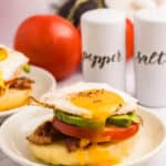 An open-faced breakfast sandwich topped with a sunny-side-up egg, avocado slices, tomato, and bacon on a toasted English muffin, served on a beige plate.
