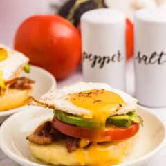 An open-faced breakfast sandwich topped with a sunny-side-up egg, avocado slices, tomato, and bacon on a toasted English muffin, served on a beige plate.