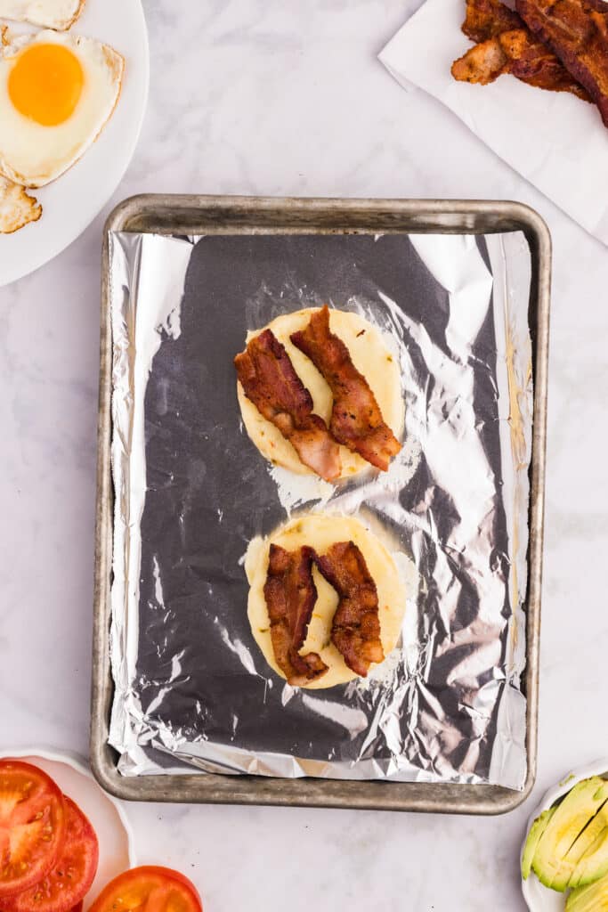 A foil-lined baking tray holds two biscuit halves topped with crispy bacon strips. Sliced tomatoes, a fried egg, avocado, and extra bacon are visible around the tray on a white marble surface.