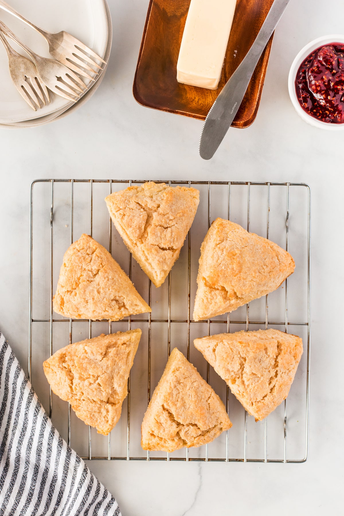 Six golden scones arranged in a circle on a cooling rack, with a plate and forks, a dish of butter with a knife, and a small bowl of jam nearby on a white surface.