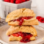 A cut scone filled with raspberry jam and cream is stacked on a plate, with fresh raspberries in a container in the background.