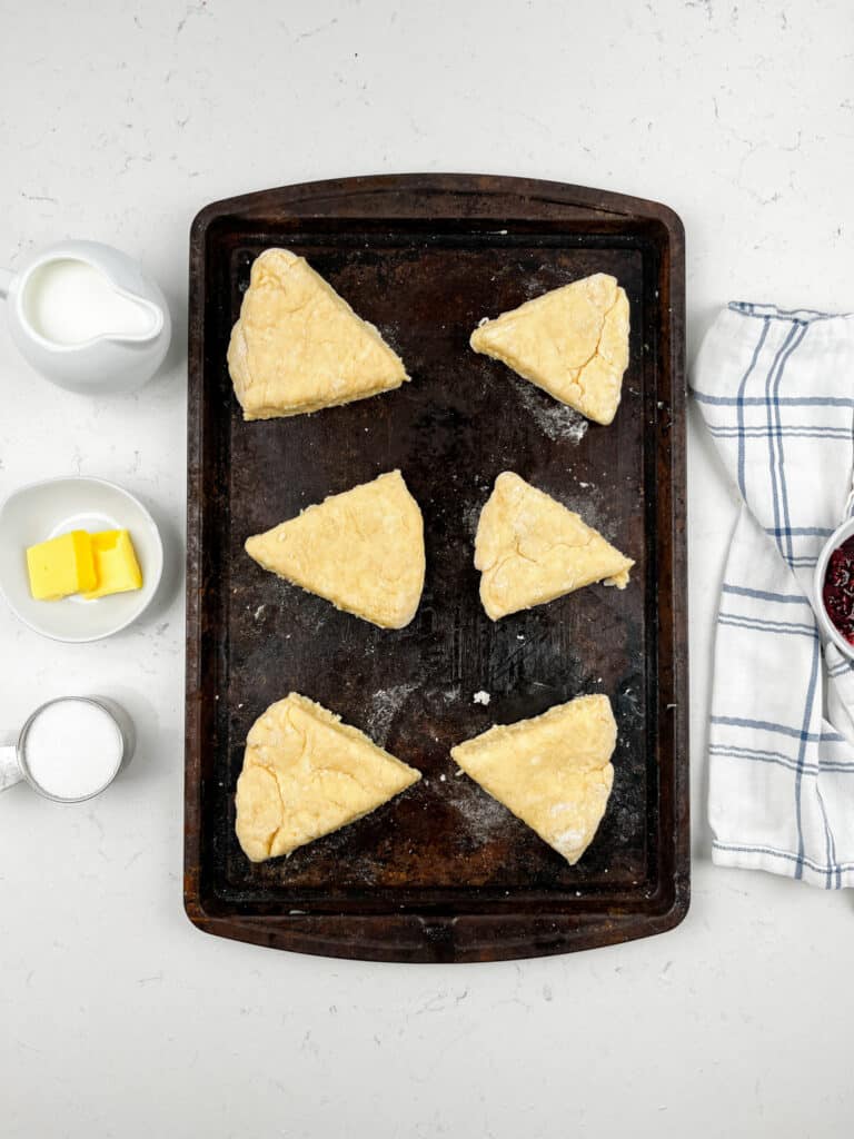 A baking tray with six unbaked scones arranged in a circle on a white surface. Nearby are small bowls of butter, milk, and sugar, a striped kitchen towel, and a bowl of jam.