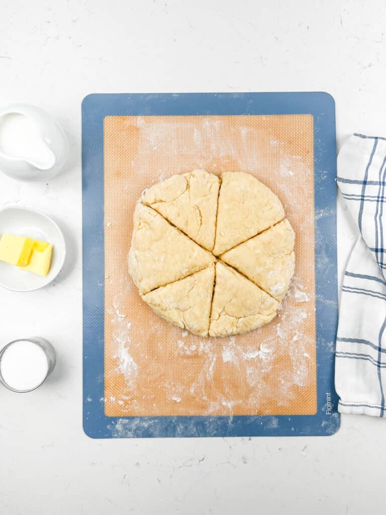 A round dough disc cut into six wedges sits on a floured baking mat. To the left are a small pitcher of milk, a dish of butter, and a bowl of sugar. A striped kitchen towel lies on the right.