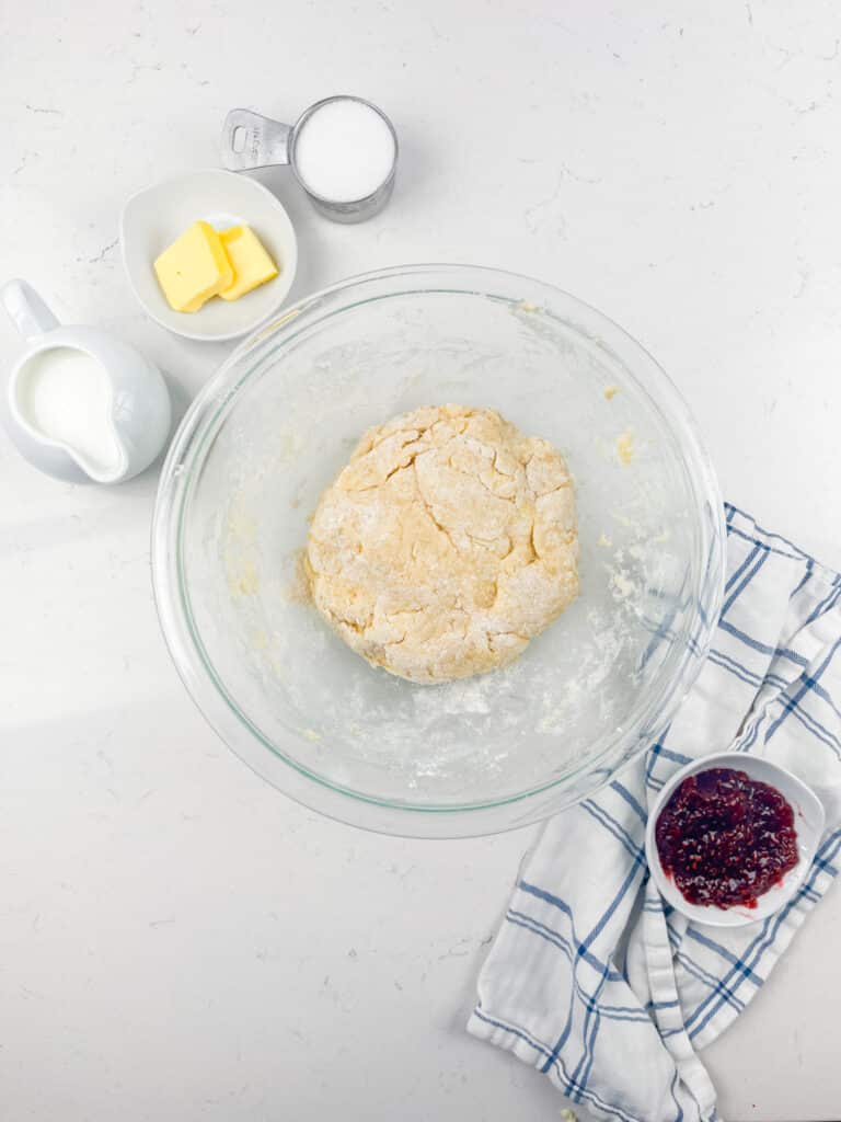 A glass bowl with dough sits on a white counter, surrounded by a small pitcher of milk, a dish of butter, a measuring cup of sugar, and a bowl of red jam on a blue and white striped cloth.