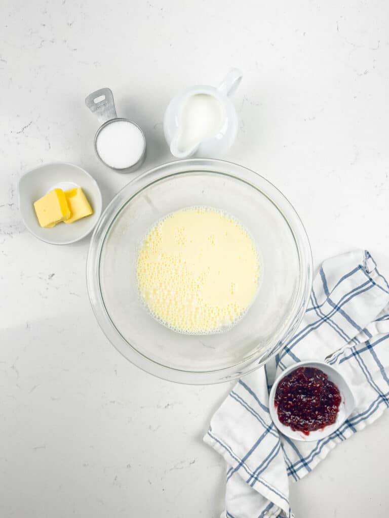 A glass bowl with a yellow mixture sits on a white countertop, surrounded by a cup of sugar, a small pitcher of milk, a bowl of butter, and a bowl of red jam on a white and blue cloth.