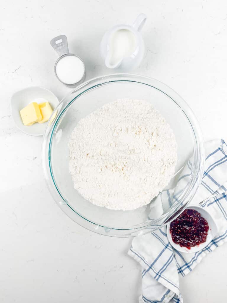 Overhead view of a glass bowl with flour, a small pitcher of milk, a cup of sugar, a dish with butter, and a bowl of jam on a white surface with a blue-striped cloth.