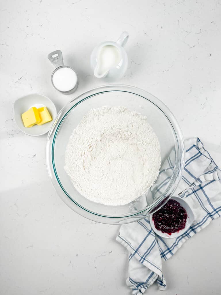 A bowl of flour sits on a white countertop, surrounded by a small pitcher of milk, a cup of sugar, a dish with butter, and a bowl of berry jam on a checkered cloth.