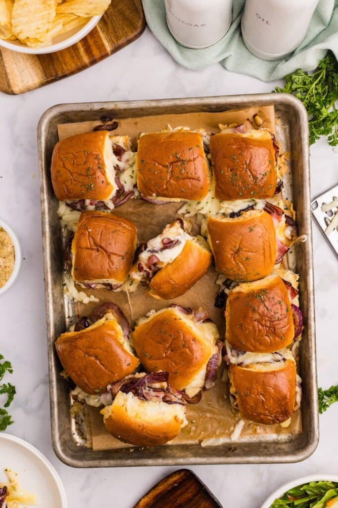 A baking tray with twelve golden-brown sliders filled with melted cheese and roast beef, set on parchment paper. The sliders are garnished with herbs. Surrounding the tray are small dishes and fresh herbs.