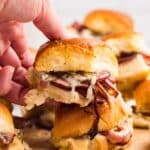 A hand lifts a slider sandwich filled with melted cheese, sliced meat, and onions from a wooden board with more sliders stacked in the background.