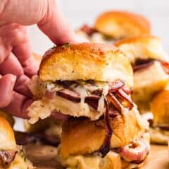 A hand lifts a slider sandwich filled with melted cheese, sliced meat, and onions from a wooden board with more sliders stacked in the background.