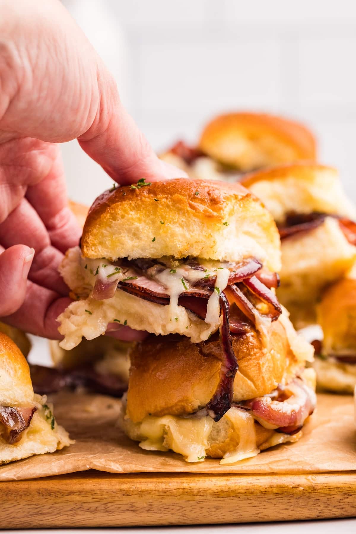 A hand lifts a slider sandwich filled with melted cheese, sliced meat, and onions from a wooden board with more sliders stacked in the background.