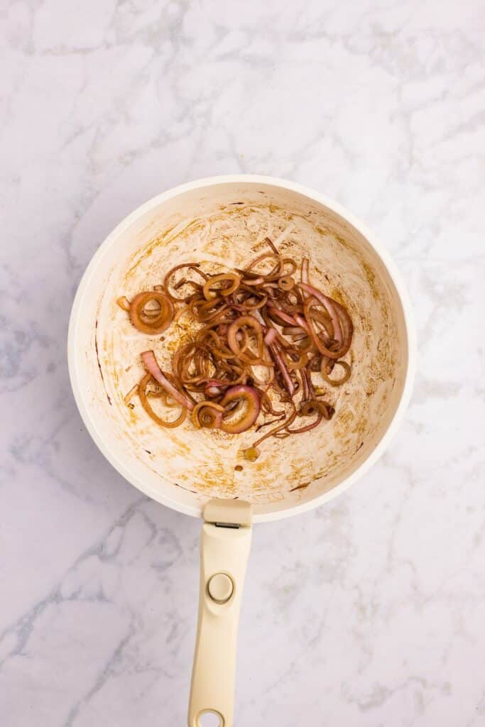 A white saucepan with sautéed sliced red onions and browned residue at the bottom, sitting on a light marble countertop.