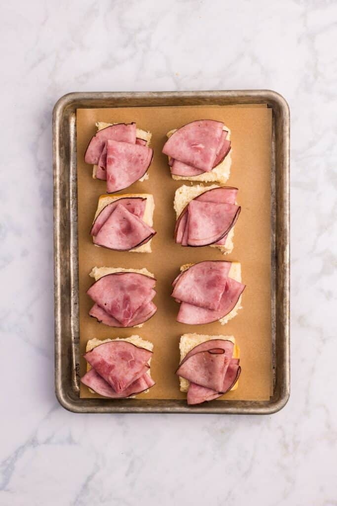 A baking tray lined with parchment paper holds eight pieces of sandwich bread, each topped with folded slices of ham. The tray is on a white marble surface.