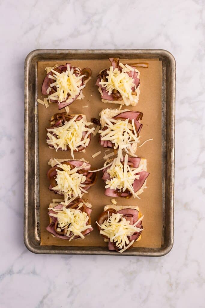 A baking tray lined with parchment paper holds eight pieces of bread topped with sliced ham, caramelized onions, and shredded cheese, arranged in two neat rows on a marble surface.