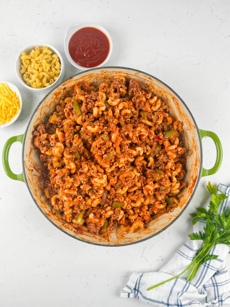 A large green pot filled with macaroni mixed with ground meat and tomato sauce is on a white surface. Nearby are bowls of shredded cheese, elbow macaroni, and tomato sauce, plus parsley and a blue-striped towel.