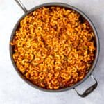 A large skillet filled with cooked elbow macaroni mixed with ground meat and tomato sauce, creating a hearty pasta dish. The skillet sits on a light gray surface.