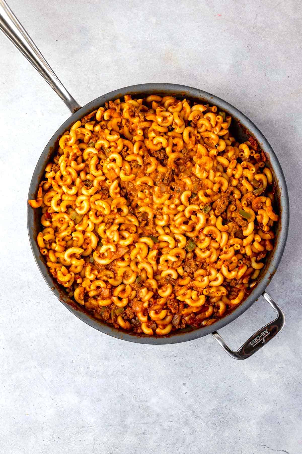 A large skillet filled with cooked elbow macaroni mixed with ground meat and tomato sauce, creating a hearty pasta dish. The skillet sits on a light gray surface.