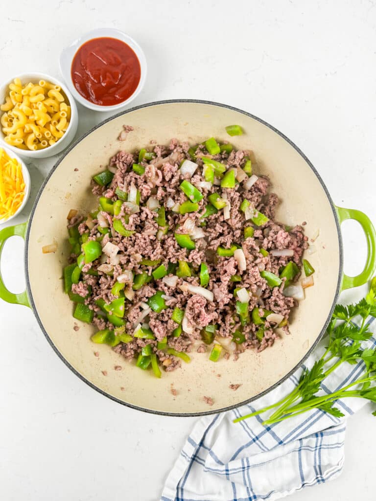 A pan filled with cooked ground beef, chopped green bell peppers, onions, and mushrooms sits on a white countertop. Surrounding the pan are small bowls of uncooked macaroni, shredded cheese, ketchup, and a bunch of parsley.