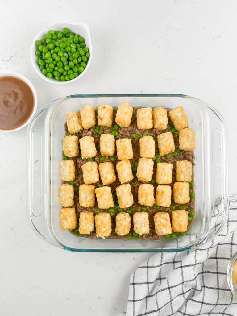 A glass baking dish filled with a layer of peas and ground meat, topped with rows of tater tots. Nearby are a bowl of peas, a bowl of brown sauce, and a striped kitchen towel on a white countertop.