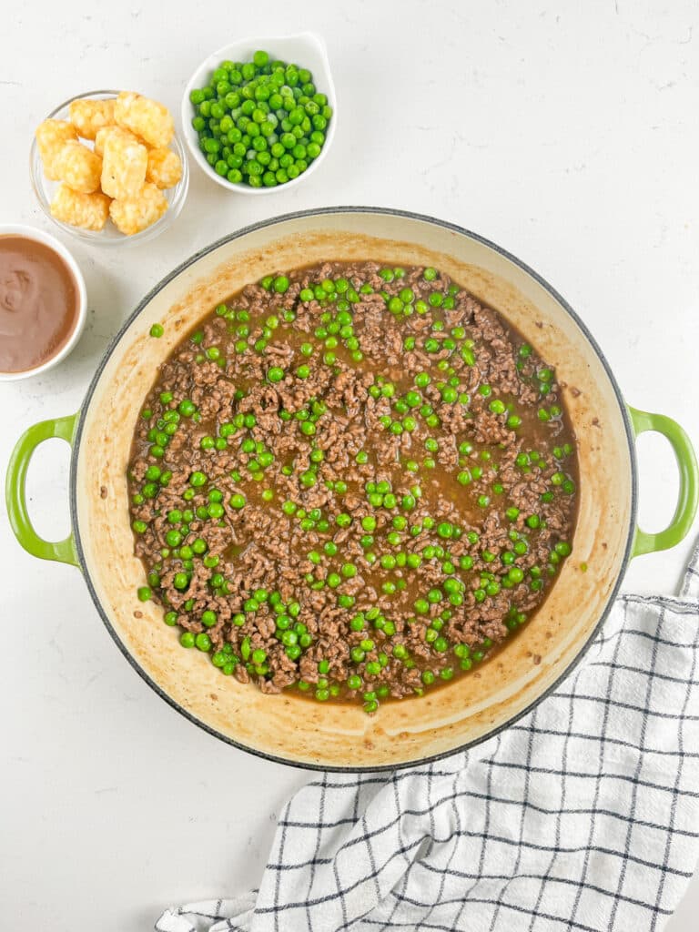 A large green pot filled with cooked ground beef and green peas in a brown sauce sits on a white surface, next to bowls of tater tots, green peas, gravy, and a checkered kitchen towel.