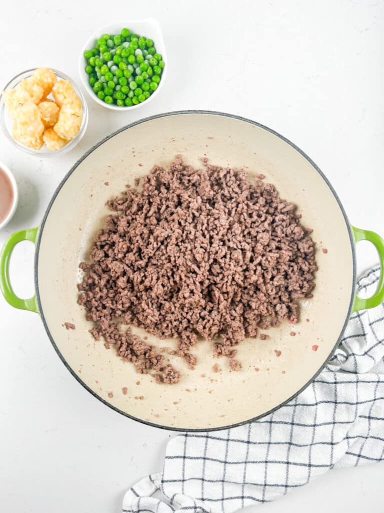 A pot with cooked ground beef sits on a white surface next to small bowls of tater tots and green peas, with a white checkered kitchen towel nearby.