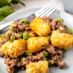 A serving of tater tot casserole with ground beef, peas, and golden tater tots sits on a gray plate with a fork beside it. Green herbs and a casserole dish are blurred in the background.