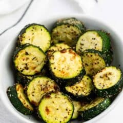 A white bowl filled with roasted zucchini slices seasoned with herbs and spices, set on a white surface with a striped cloth partially visible in the background.