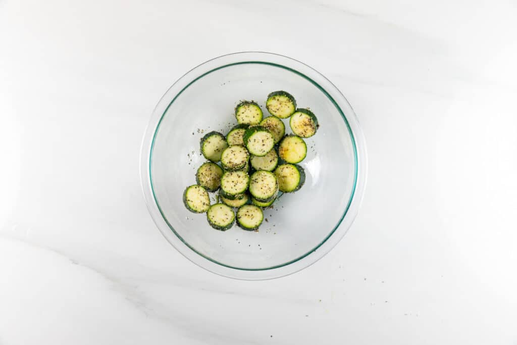 A glass bowl filled with sliced cucumbers seasoned with herbs and black pepper, placed on a white marble surface.