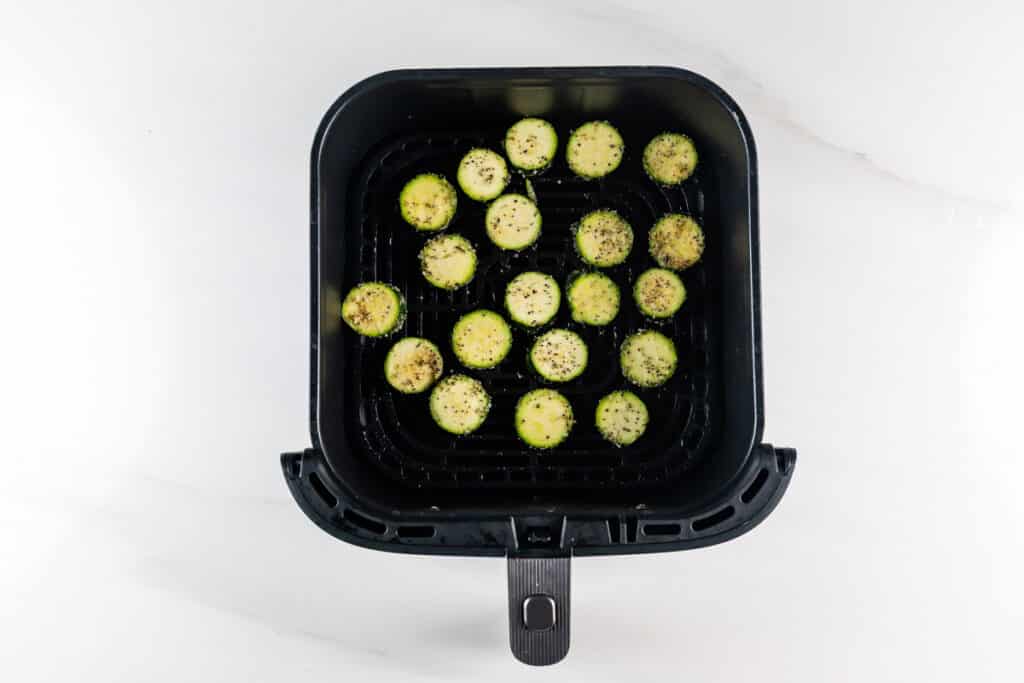 Sliced zucchini rounds arranged in a single layer inside an air fryer basket, seasoned with black pepper and ready to cook. The air fryer is placed on a white surface.