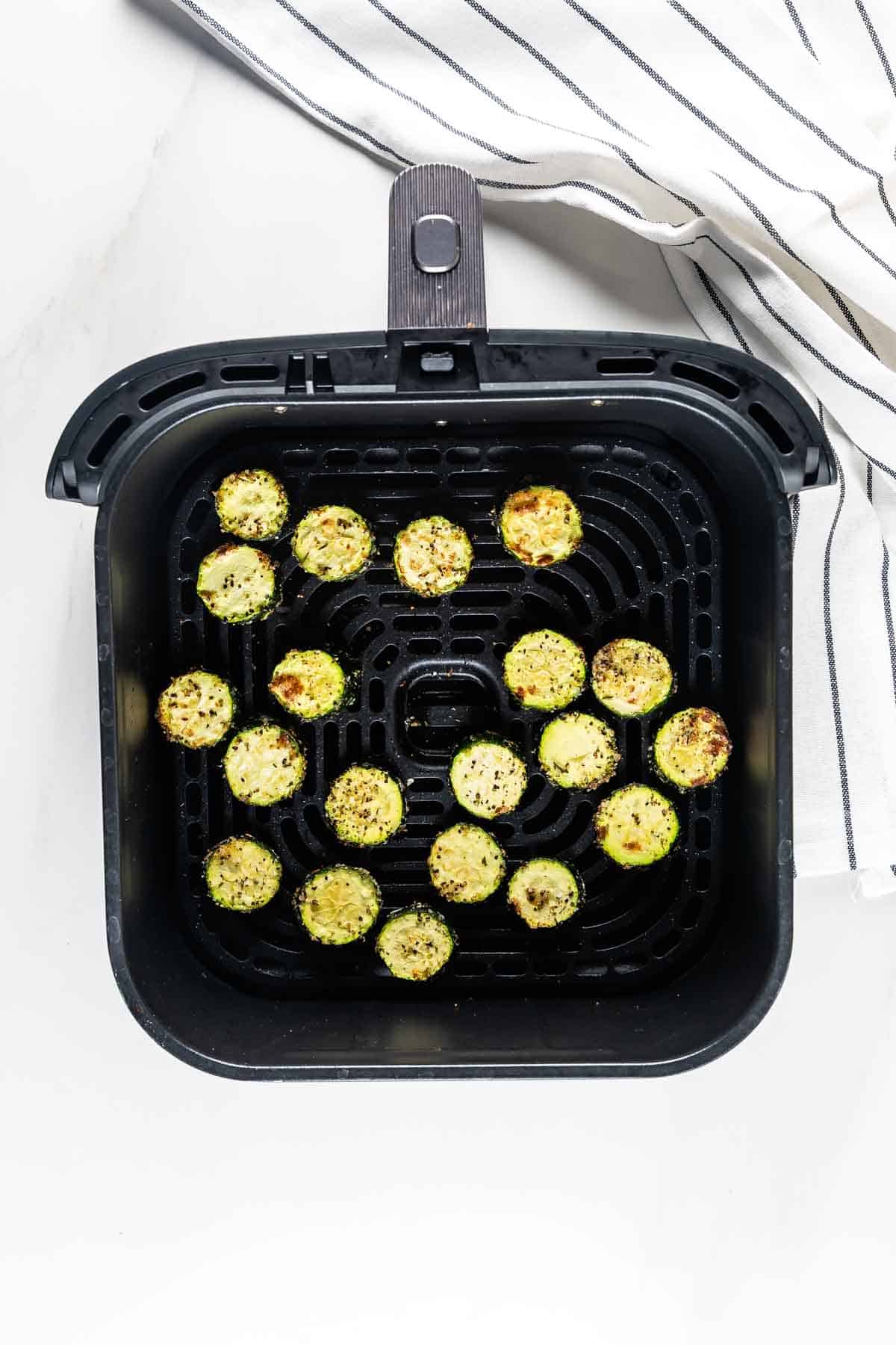 Sliced zucchini rounds, seasoned and cooked, arranged in a single layer inside an air fryer basket. A striped kitchen towel is partially visible next to the basket.