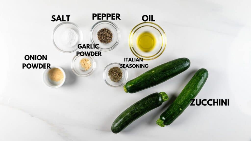 Three whole zucchinis beside small bowls containing salt, pepper, oil, onion powder, garlic powder, and Italian seasoning, all labeled, on a white surface.