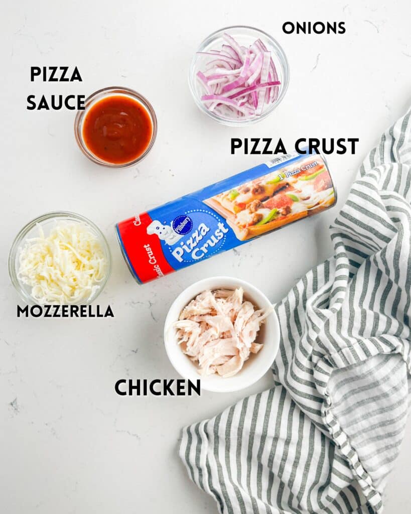 A flat lay image of pizza sauce, shredded mozzarella, sliced red onions, shredded chicken, and a tube of pizza crust on a white surface with a striped kitchen towel, each ingredient labeled in black text.