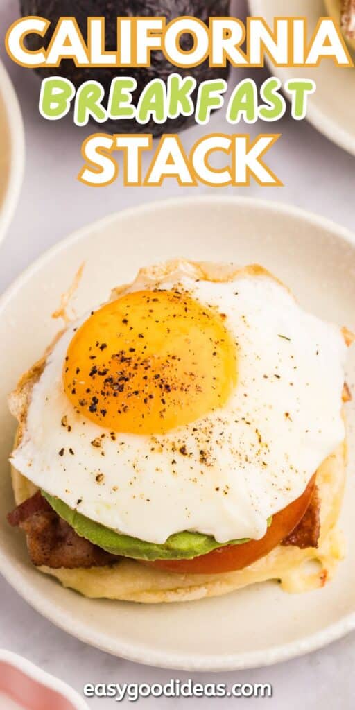 An open-faced breakfast sandwich topped with a sunny-side-up egg, avocado slices, tomato, and bacon on a toasted English muffin, served on a beige plate. Text above reads California Breakfast Stack.
