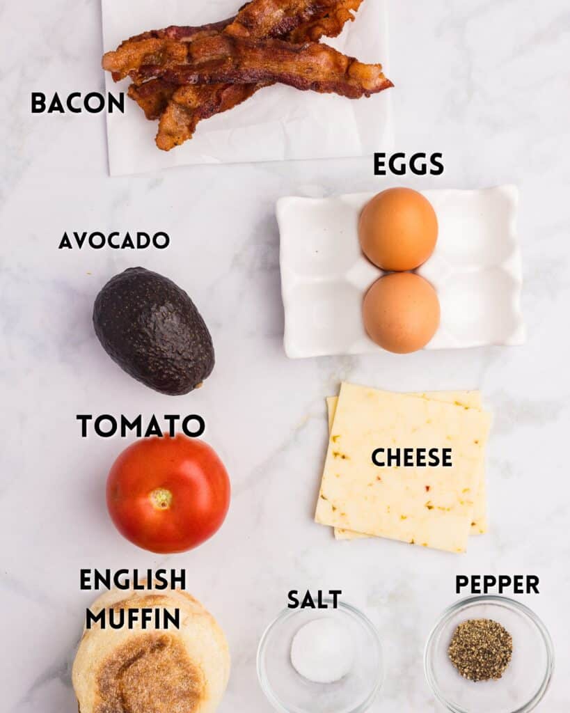 Flat lay of breakfast sandwich ingredients on a white surface: two strips of bacon, two eggs, an avocado, a tomato, two slices of cheese, an English muffin, a small pile of salt, and a small pile of pepper. Labels identify each item.