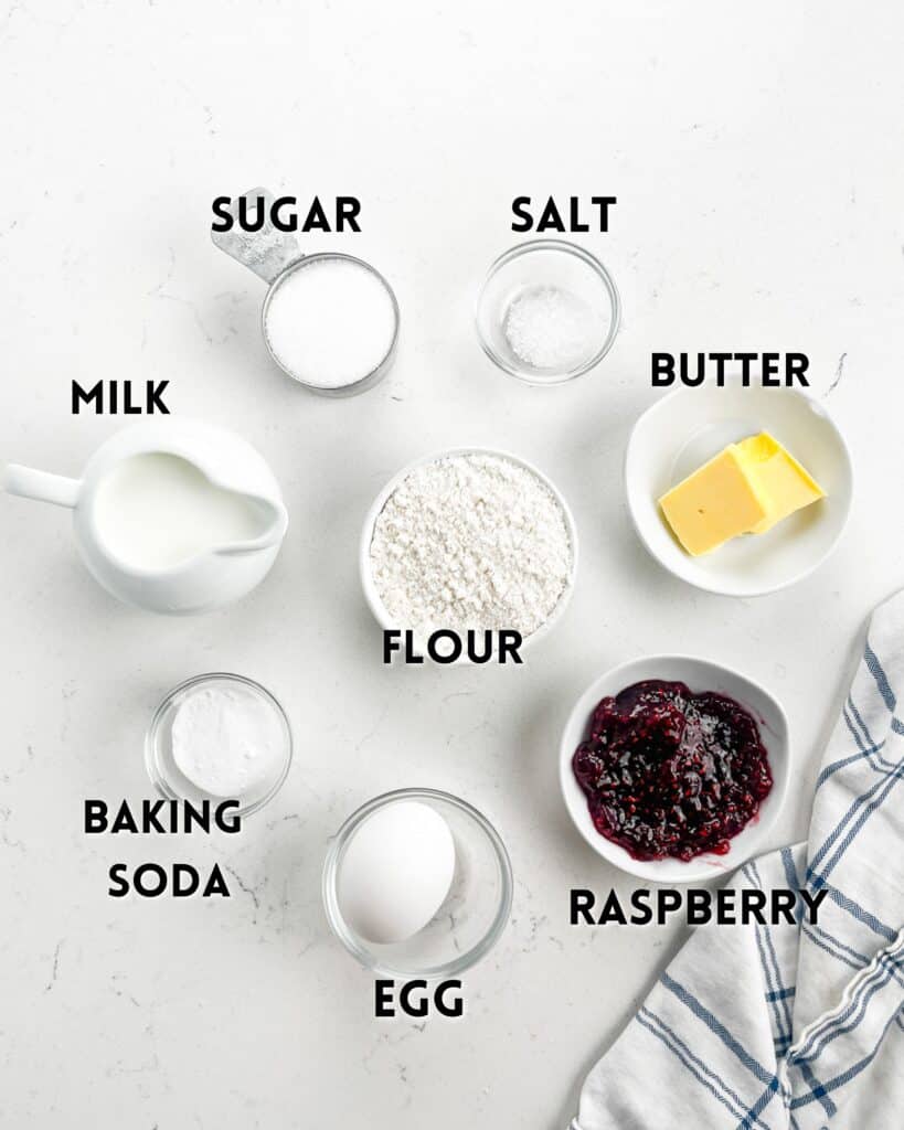 Seven labeled ingredients—sugar, salt, butter, flour, baking soda, egg, raspberry, and milk—are arranged on a white surface next to a striped kitchen towel.
