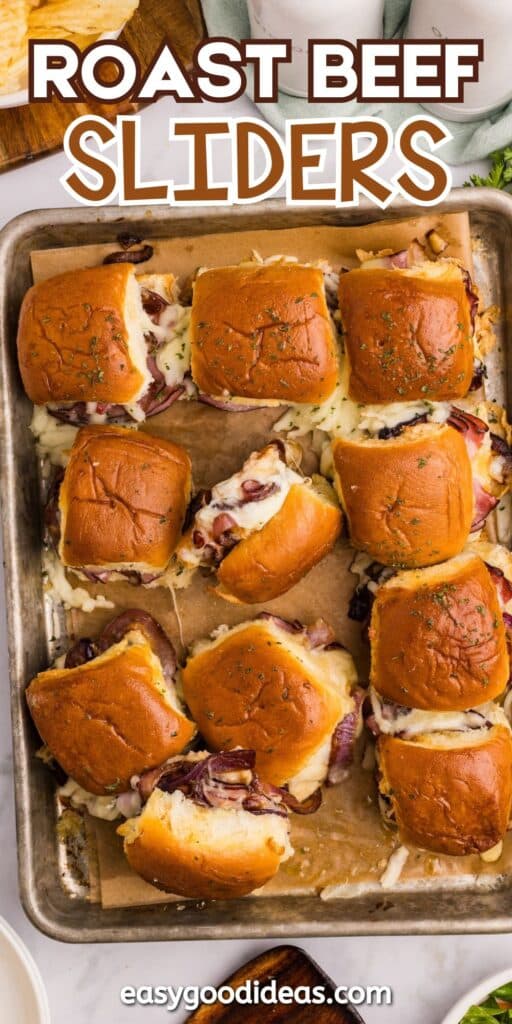 A baking sheet lined with parchment paper holds nine roast beef sliders on golden buns, topped with melted cheese and herbs. Some cheese and filling have melted out onto the tray. Text reads Roast Beef Sliders and easygoodideas.com.