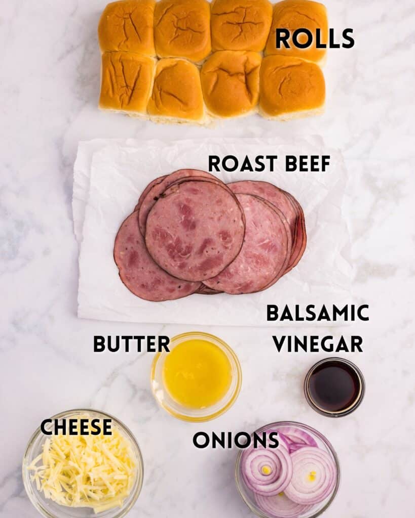 A flat lay of sandwich ingredients labeled: soft rolls, sliced roast beef, shredded cheese, sliced onions, melted butter in a bowl, and balsamic vinegar in a small dish on a marble surface.