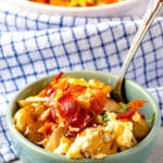 A bowl filled with cheesy potato casserole topped with crispy bacon and herbs, with a spoon inside. A larger dish of the same casserole is in the background, set on a blue-and-white checkered cloth.