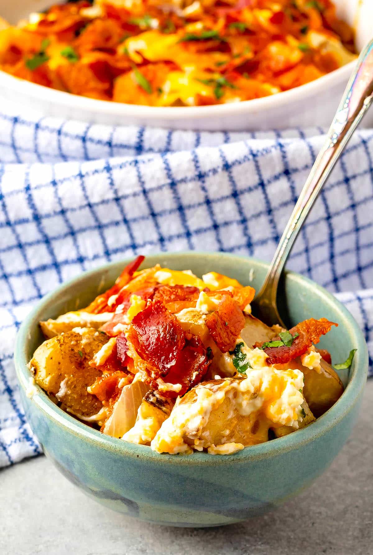 A bowl filled with cheesy potato casserole topped with crispy bacon and herbs, with a spoon inside. A larger dish of the same casserole is in the background, set on a blue-and-white checkered cloth.