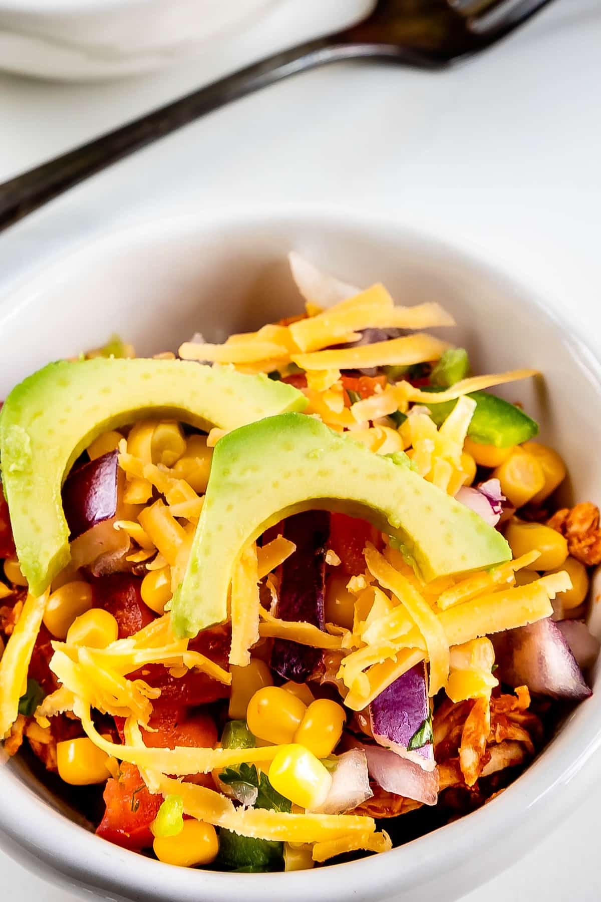 A close-up of a bowl filled with sliced avocado, shredded cheddar cheese, corn, diced red onion, tomatoes, and lettuce, creating a colorful and fresh salad. A spoon is visible in the background.